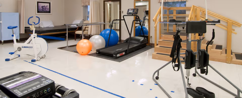 Rehabilitation gym with a treadmill, exercise bike, therapy balls, parallel bars and practice stairs.