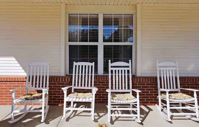 Four white rocking chairs with patterned cushions sit on a porch in front of a window and brick facade.