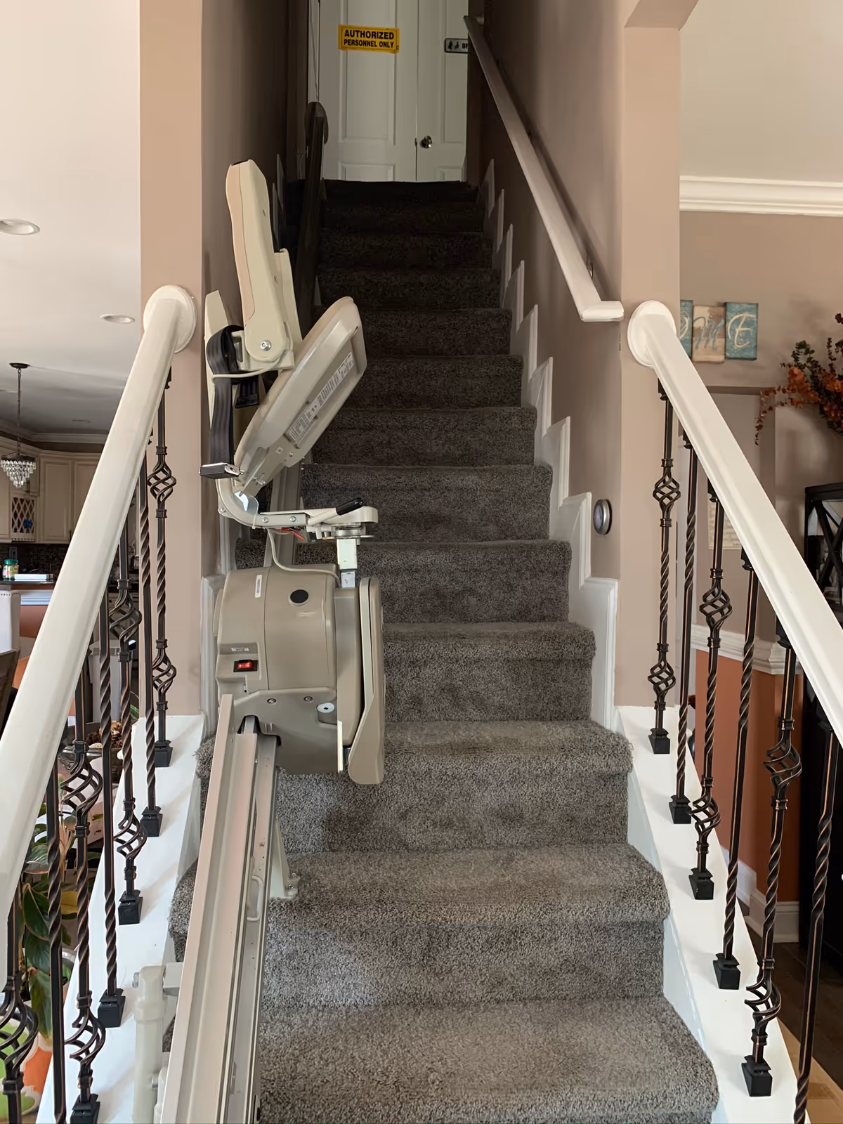 Carpeted staircase with a stair lift installed on the left side. The stair lift chair is folded up. The staircase has white railings with decorative black balusters. At the top of the stairs, there is a white door with a yellow sign that reads 'AUTHORIZED PERSONNEL ONLY'. Part of a kitchen and living area is visible to the left and right of the staircase.