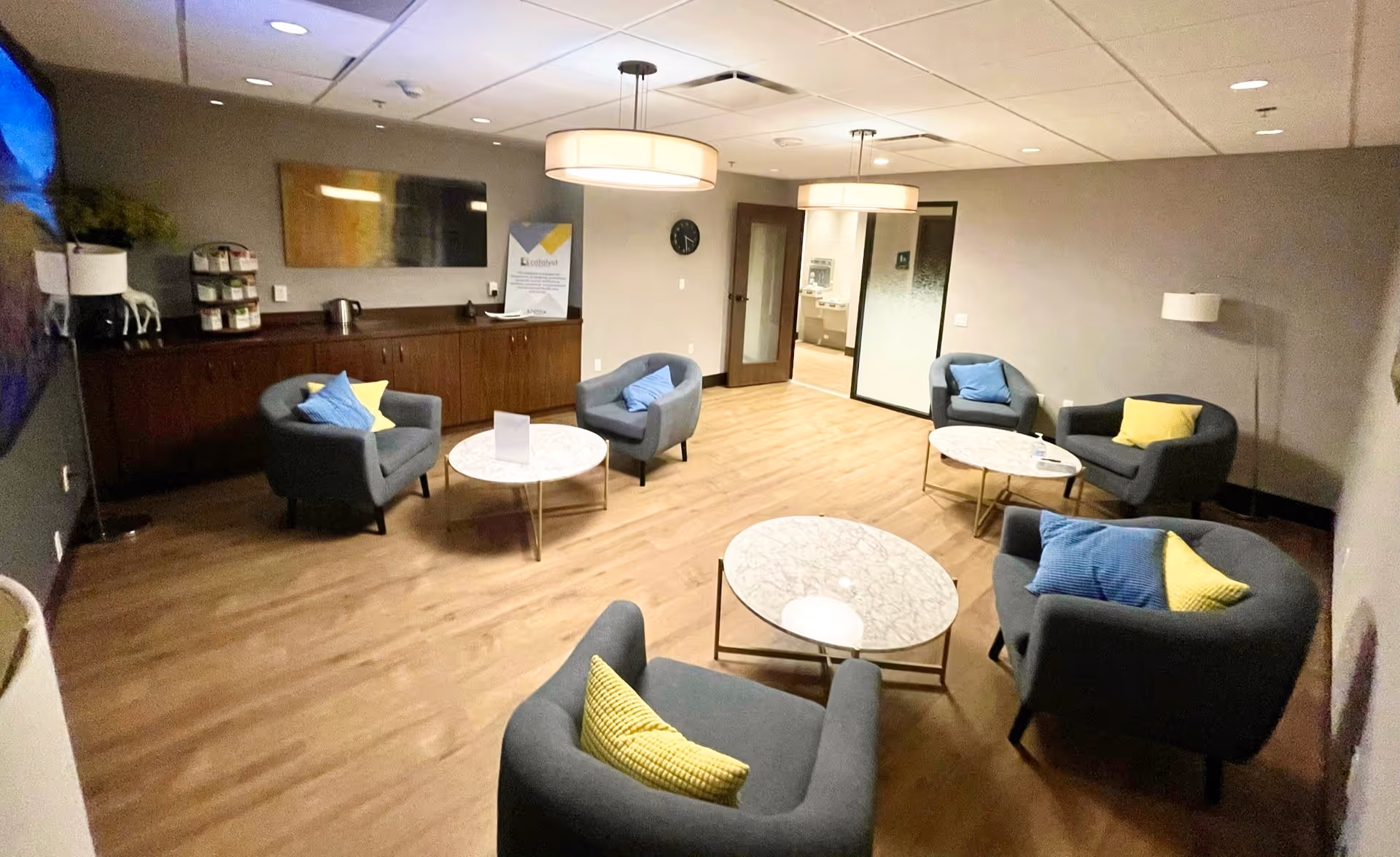 A modern lounge area with six gray armchairs, each adorned with blue and yellow pillows, arranged around three round marble-top coffee tables. The room has light wooden flooring, neutral-colored walls, two large ceiling lights, a wall-mounted TV, and a wooden cabinet along one wall. There is a clock on the wall and a door leading to another room in the background.