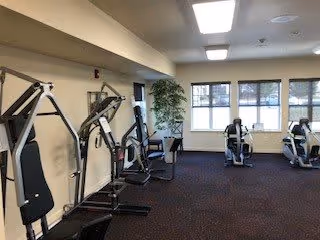 A fitness room with exercise equipment including weight machines and stationary bikes. The room has carpeted flooring, large windows letting in natural light, and a potted plant near the windows.