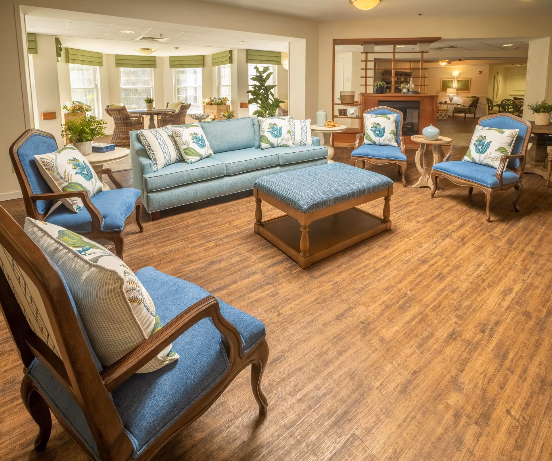 A bright and spacious living room area with wooden flooring, featuring a light blue sofa, four blue upholstered armchairs with floral cushions, a matching blue ottoman, and small round wooden side tables. Large windows with green valances allow natural light to fill the room, and there are plants and decorative items placed around the space.