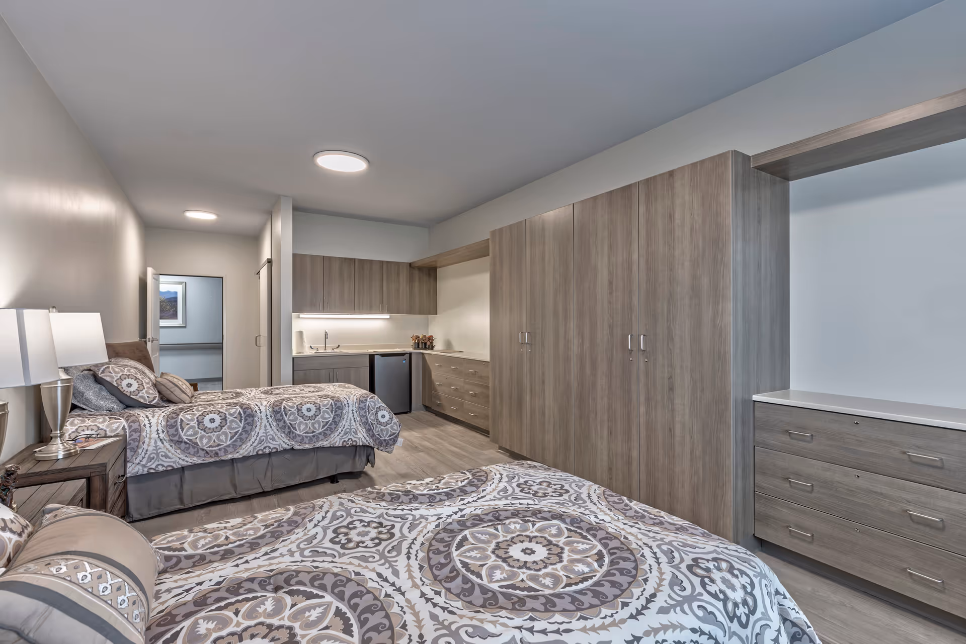 Spacious senior living bedroom with two beds, bedside tables, built-in wood cabinets and a small kitchenette area.