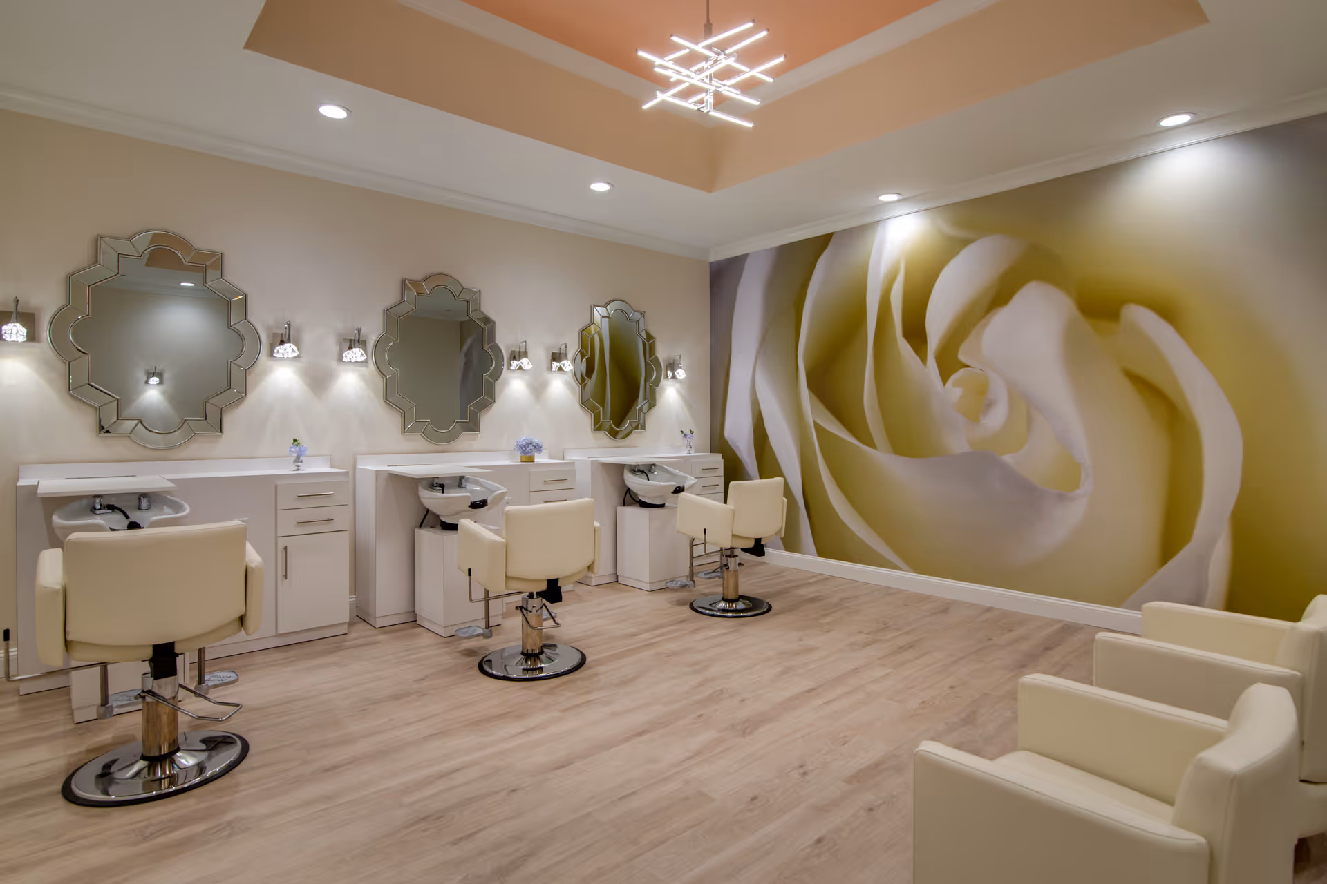 A modern salon area with three white styling chairs in front of three decorative mirrors mounted on a beige wall. Each station has a white cabinet with a sink. The floor is light wood, and the opposite wall features a large mural of a white rose. The ceiling has recessed lighting and a contemporary chandelier.