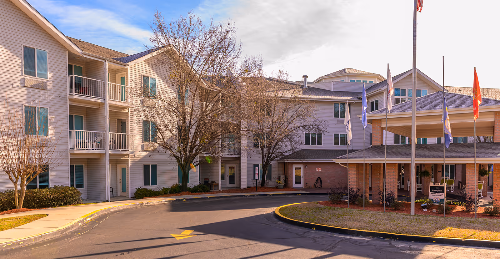 Front entrance and facade of a multi-story senior living facility with balconies, flagpoles, and a circular drive-up.