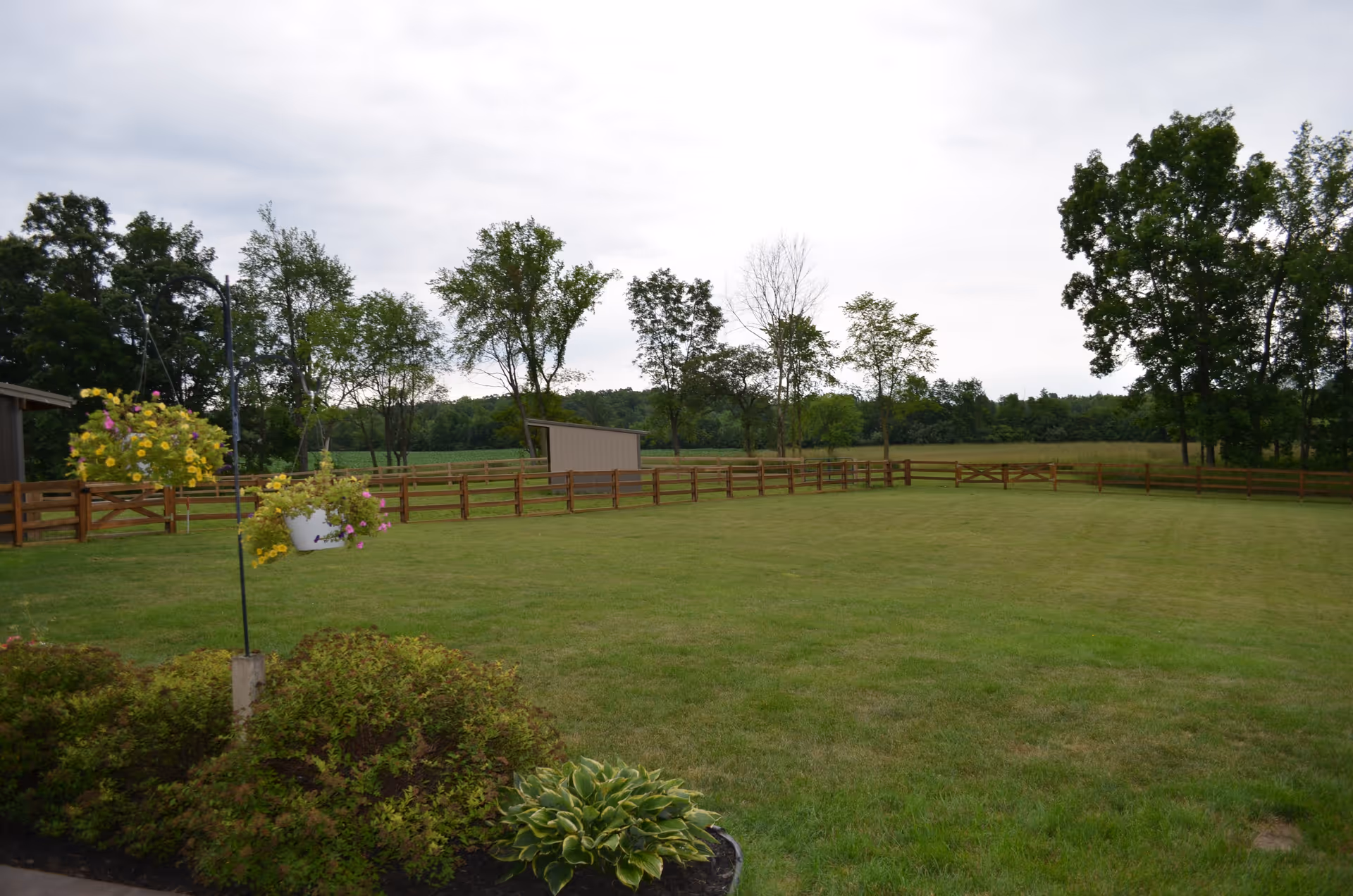 Well-maintained grassy yard with a wooden fence, hanging flower baskets, and trees under a cloudy sky.