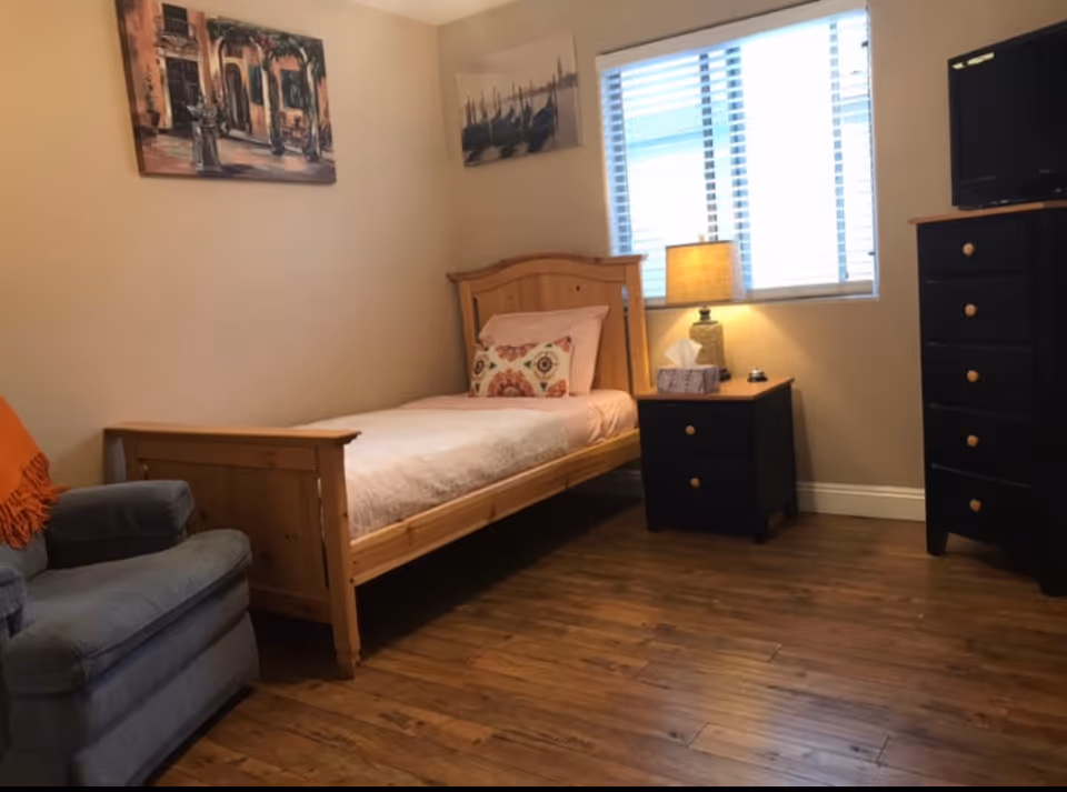 A cozy bedroom with a wooden single bed featuring a white bedspread and decorative pillows. Next to the bed is a black nightstand with a lamp, tissue box, and a small item on it. There is a window with blinds letting in natural light. A blue armchair with an orange throw blanket is positioned near the bed. On the right side, there is a tall black dresser with wooden knobs and a small TV on top. The walls are decorated with two paintings, and the floor is wooden.