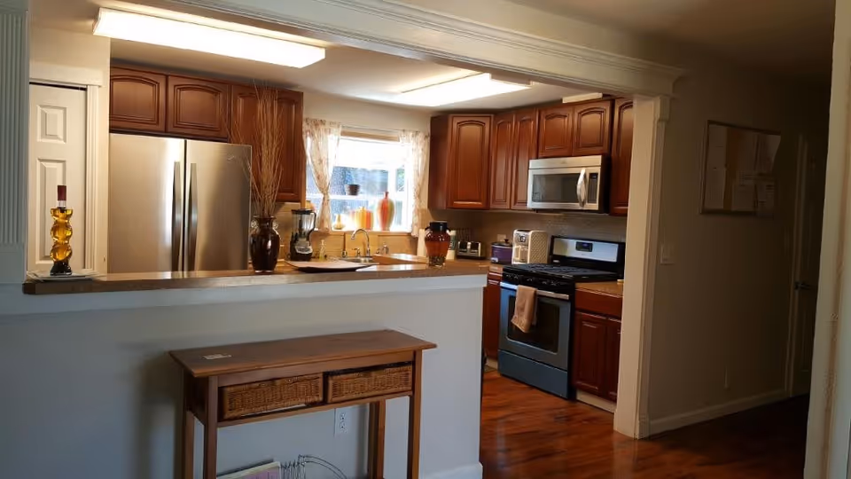 A well-lit kitchen with wooden cabinets, a stainless steel refrigerator, a stove with an oven, a microwave, and a countertop with decorative vases. There is a small wooden table with two wicker baskets underneath in the foreground. A window above the sink lets in natural light.