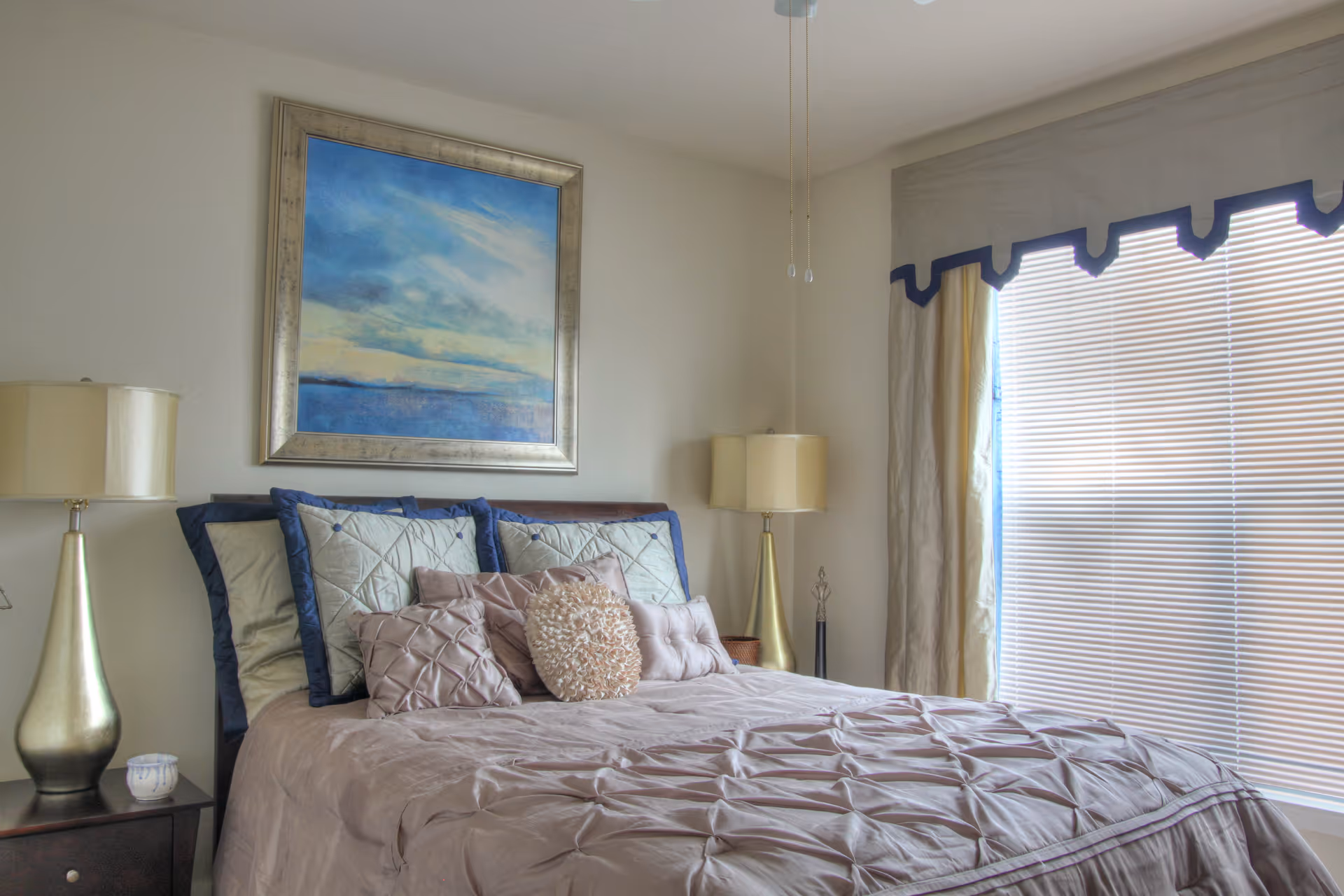 Well-lit bedroom with a neatly made bed topped with decorative pillows, bedside lamps, framed artwork, and a window with blinds and a valance.