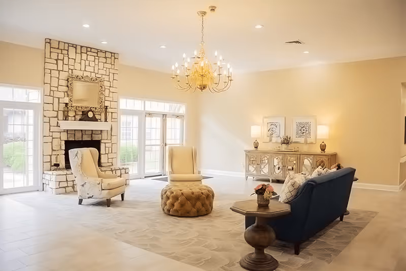 A bright and spacious living room with a stone fireplace, two beige armchairs, a tufted round ottoman, a dark blue sofa with patterned pillows, a wooden side table with a flower arrangement, and a decorative console table with lamps and framed artwork on the wall.