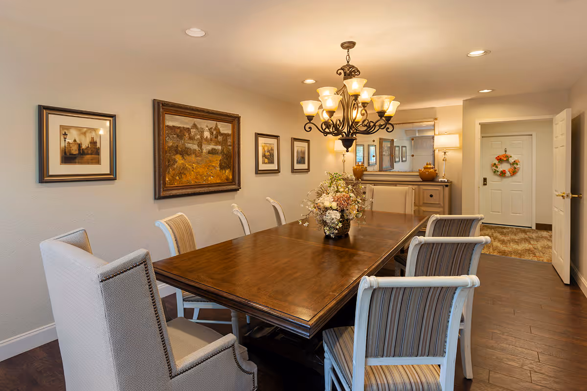 Formal dining room with a long wooden table, upholstered chairs, a central floral arrangement, chandelier, and framed wall art.