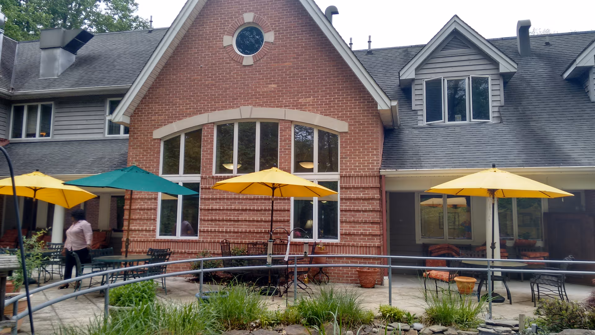 Outdoor patio area of a senior living facility with several tables and chairs under yellow and green umbrellas. The building has a brick and siding exterior with large windows and a sloped roof. There is a metal railing around the patio and some greenery in the foreground. A person is walking on the patio near one of the tables.