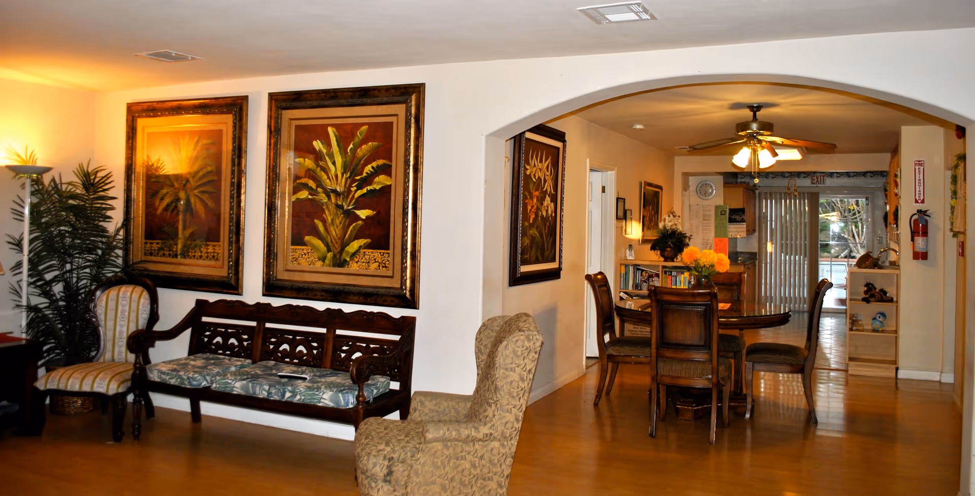 Interior view of a senior living facility common area with wooden flooring, a wooden bench with floral cushions, two armchairs, and framed botanical artwork on the walls. In the background, there is a dining area with a round wooden table and chairs, a ceiling fan with lights, and a sliding glass door leading outside.