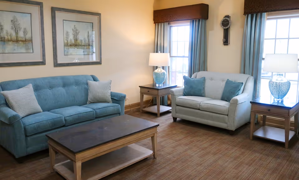 A cozy living room with two sofas, one teal and one light gray, each with matching throw pillows. There are two wooden side tables with blue and white lamps, a wooden coffee table in the center, two windows with blue curtains, and two framed landscape paintings on the wall.