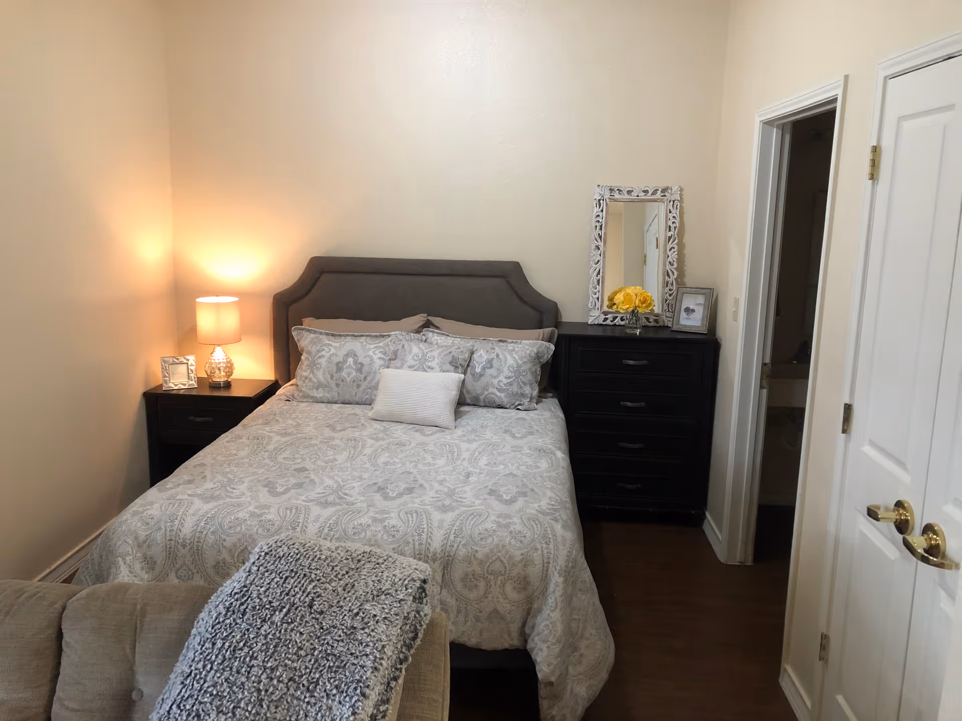 A cozy bedroom with a neatly made bed featuring patterned bedding and multiple pillows. To the left of the bed is a nightstand with a lit lamp and a small picture frame. To the right is a black dresser with a decorative mirror, a vase of yellow flowers, and a framed photo. The room has beige walls and wooden flooring, with a partially visible armchair in the foreground and a doorway leading to another room on the right.