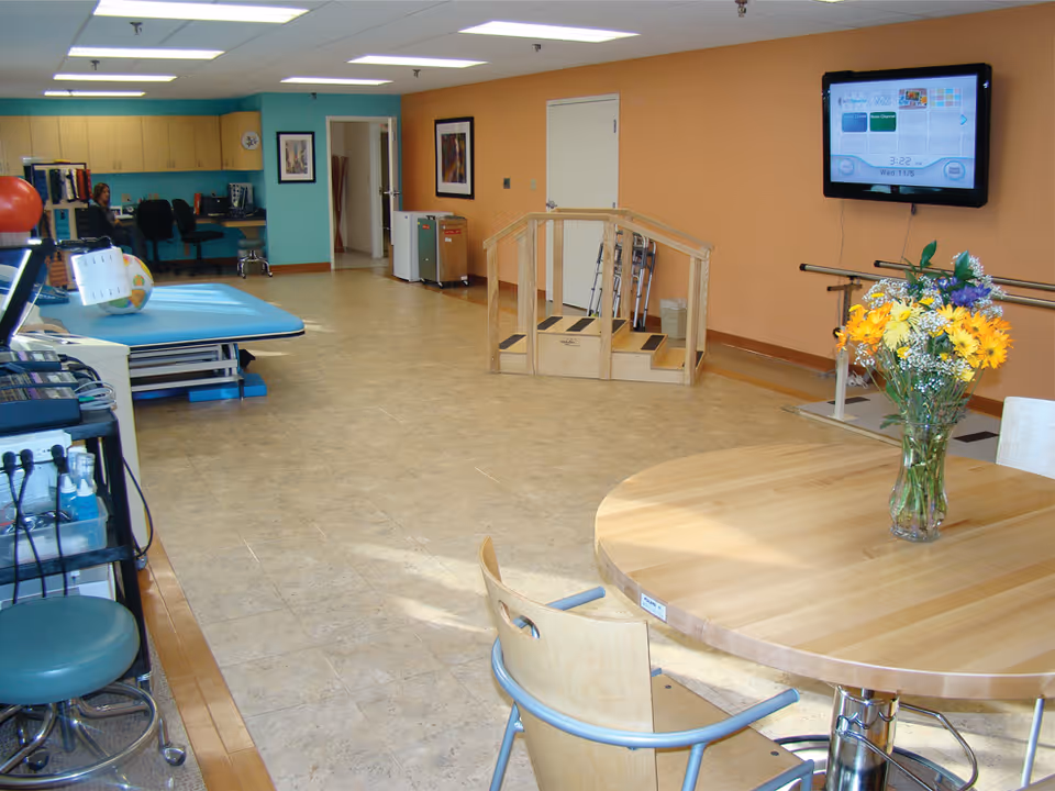 A rehabilitation therapy room with a wooden table and chairs in the foreground, a vase of yellow and purple flowers on the table, a small wooden staircase with handrails, parallel bars for walking exercises, a blue therapy table, and a wall-mounted TV screen displaying a menu. The room has a beige tiled floor and walls painted in orange and teal colors. A person is seated at a desk with cabinets in the background.