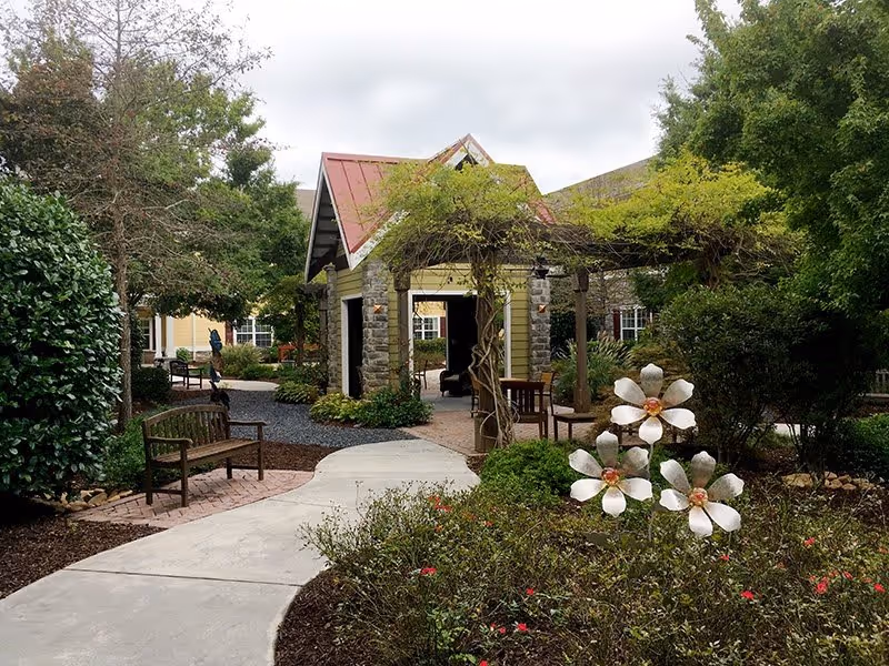 A scenic view of the entrance pathway to Park Springs, featuring a stone structure with a red roof, surrounded by lush greenery and blooming flowers.