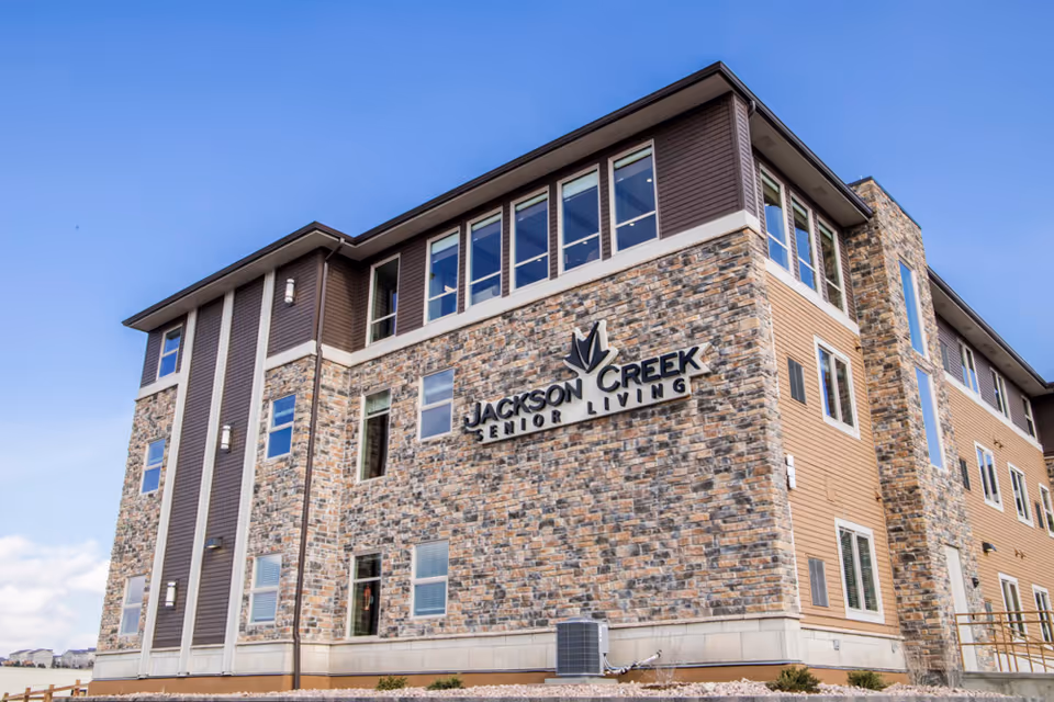 Exterior view of a multi-story building with stone and siding facade under a clear blue sky. The building has many windows and a sign that reads 'Jackson Creek Senior Living'.