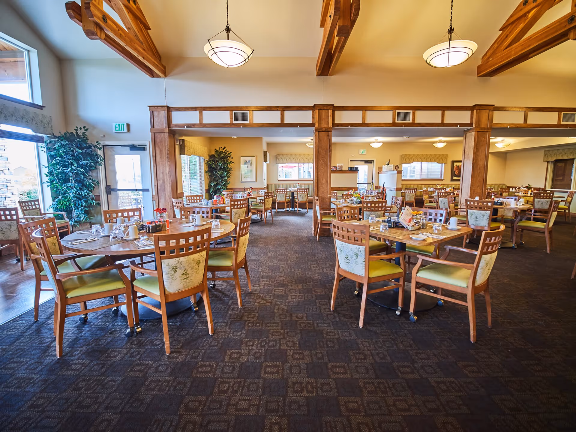 A spacious dining room with multiple wooden tables and chairs arranged neatly. The room features wooden beams on the ceiling, large windows allowing natural light, and decorative plants near the entrance. Tables are set with cups, glasses, and condiments, creating a welcoming atmosphere.