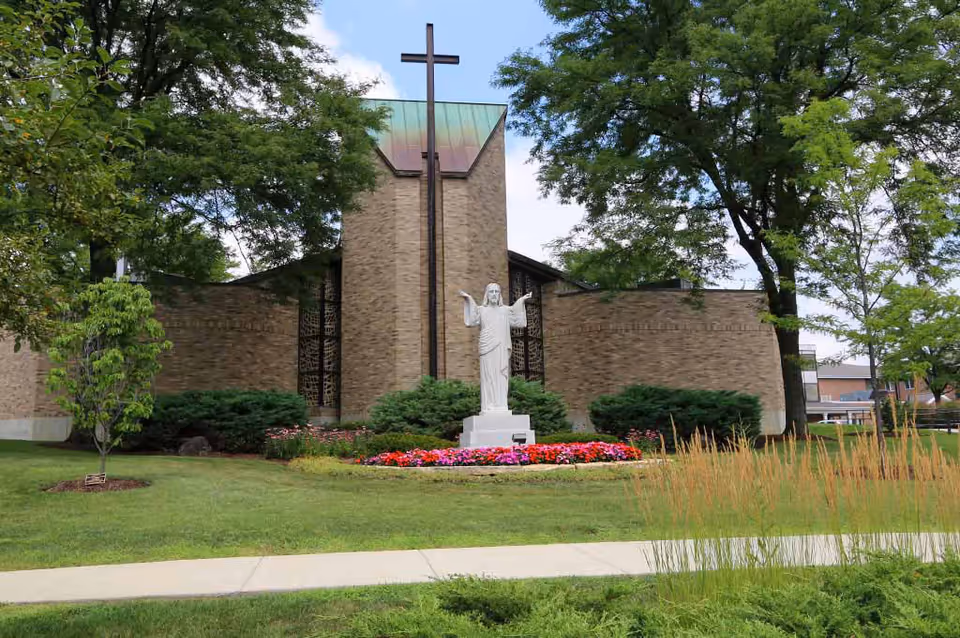 Brick building front with a tall cross and a white statue of Jesus surrounded by flowers, lawn, and trees.