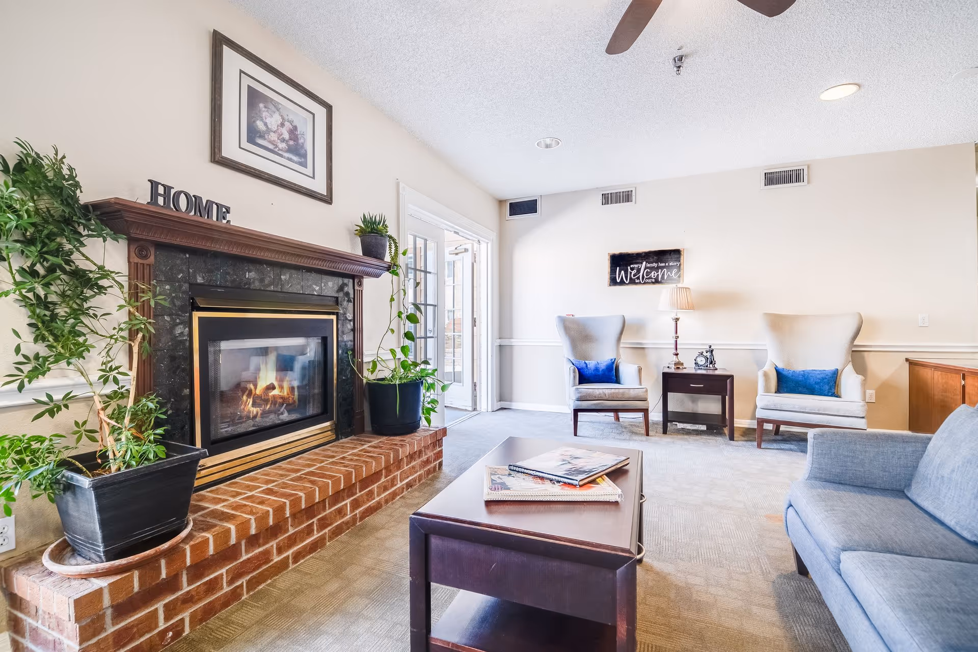 A cozy living room with a lit fireplace surrounded by a brick hearth and dark wood mantle. Two potted plants sit on the hearth. Above the fireplace is a framed floral painting and decorative letters spelling 'HOME'. The room has two beige armchairs with blue cushions, a small dark wood side table with a lamp and clock, and a blue sofa. A dark wood coffee table with magazines is in the center. A door with glass panes leads outside, and a ceiling fan is visible above.