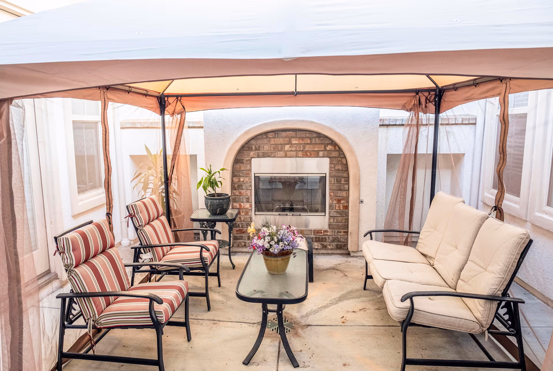 Outdoor patio area with a canopy overhead, featuring two striped cushioned chairs, a beige cushioned loveseat, a glass-top coffee table with a flower pot, and a small side table with a potted plant. A brick and stucco fireplace is built into the wall at the back.