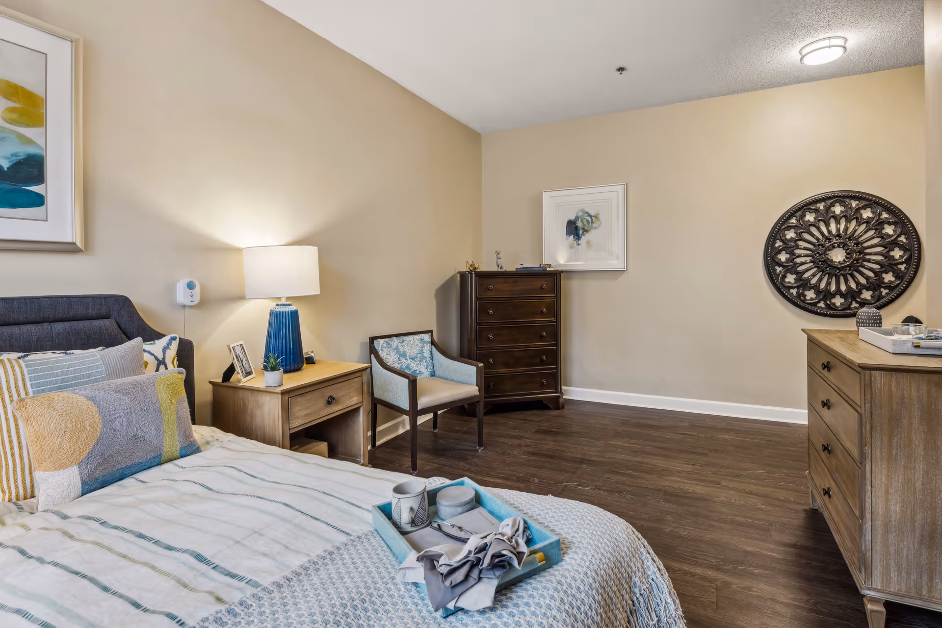 A cozy bedroom with a neatly made bed featuring multiple pillows and a tray with a cup and napkin. There is a wooden nightstand with a blue lamp, a small plant, and framed photos. A wooden chair with light blue upholstery is placed next to a dark wooden chest of drawers. On the opposite side, there is a wooden dresser with decorative items and wall art, including a framed abstract painting and a large circular ornamental piece. The room has beige walls and dark wood flooring.