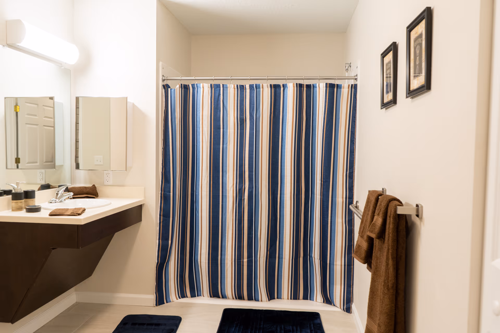 A clean bathroom with a blue, white, and beige striped shower curtain, a floating dark wood vanity with a white countertop and sink, a large mirror with a light above it, brown towels hanging on a towel rack, two framed pictures on the wall, and two dark blue bath mats on the floor.
