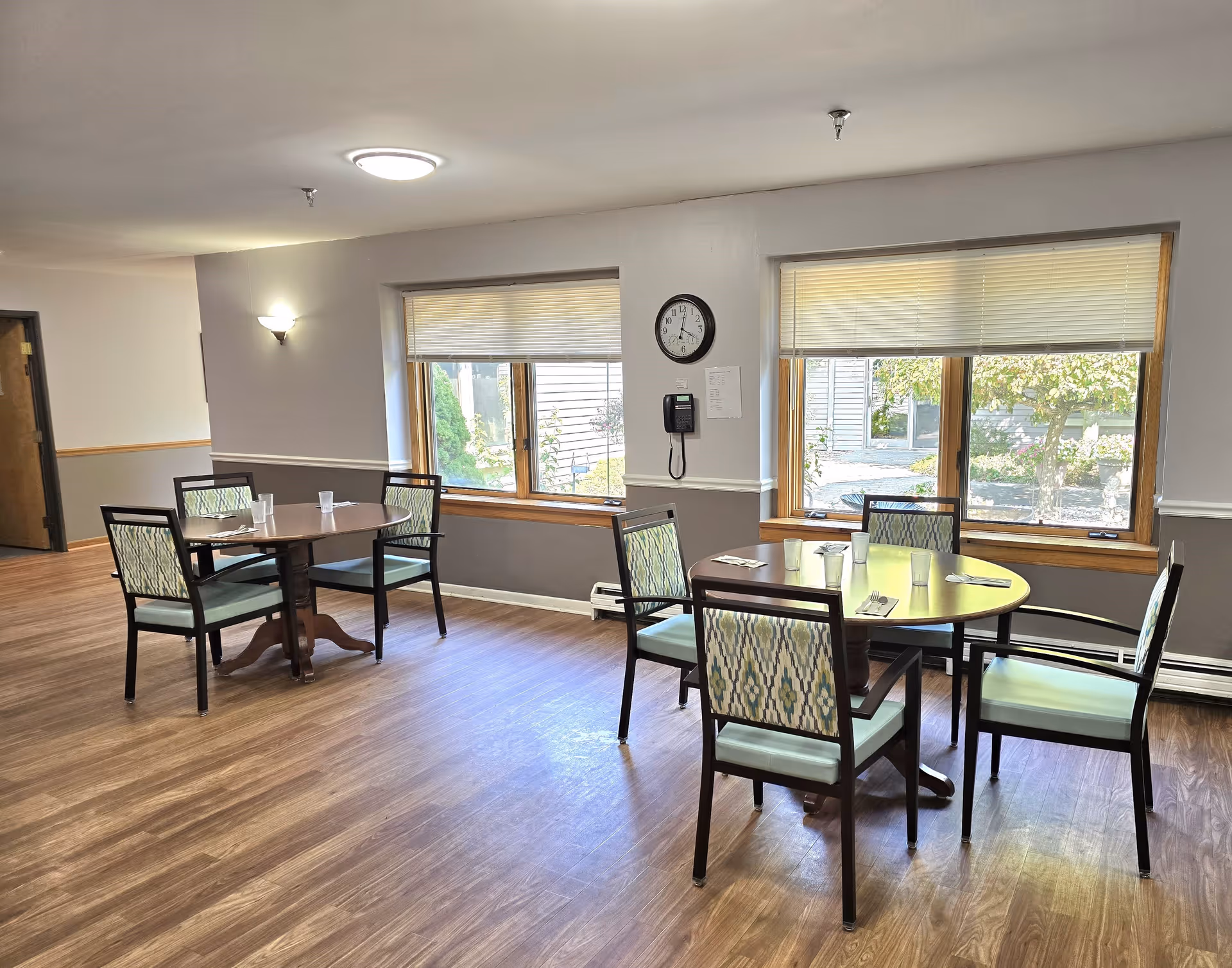 A bright dining area with two round tables, each surrounded by four chairs with patterned backs and light blue seats. The room has large windows with blinds partially drawn, allowing natural light to illuminate the space. The walls are painted gray with white trim, and there is a clock and a wall-mounted phone between the windows. The floor is wood with a light finish.
