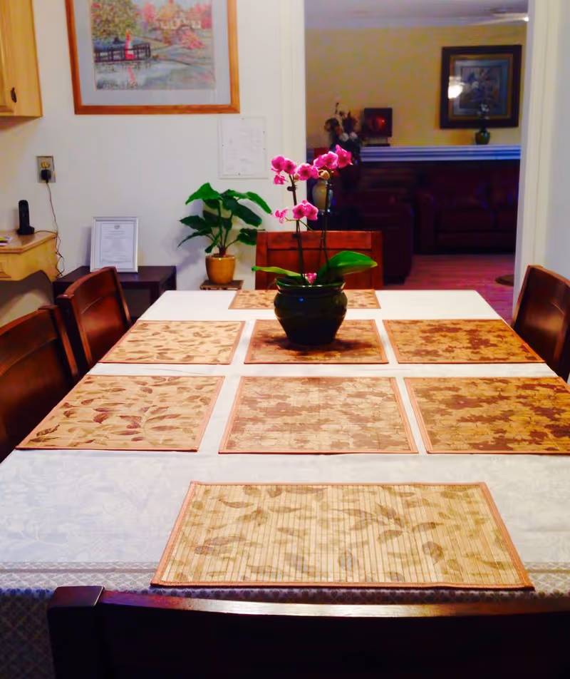 A dining table set with placemats and a potted pink orchid as a centerpiece, surrounded by chairs with a view into a living room.