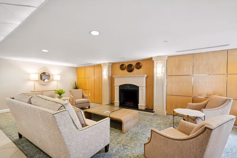A cozy living room area with a beige sofa, two beige armchairs, a wooden coffee table, and a fireplace with decorative plates above it. The room has soft lighting, a patterned carpet, and wooden paneling on the walls.