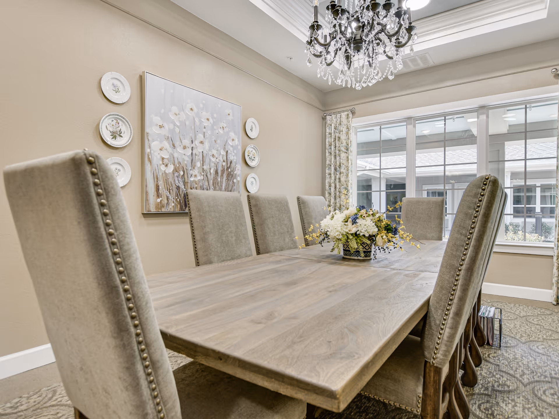 A dining room with a long wooden table surrounded by eight upholstered chairs with nailhead trim. A floral centerpiece sits in the middle of the table. The room has beige walls decorated with a large floral painting and decorative plates. A chandelier hangs from the ceiling, and large windows with floral curtains let in natural light.