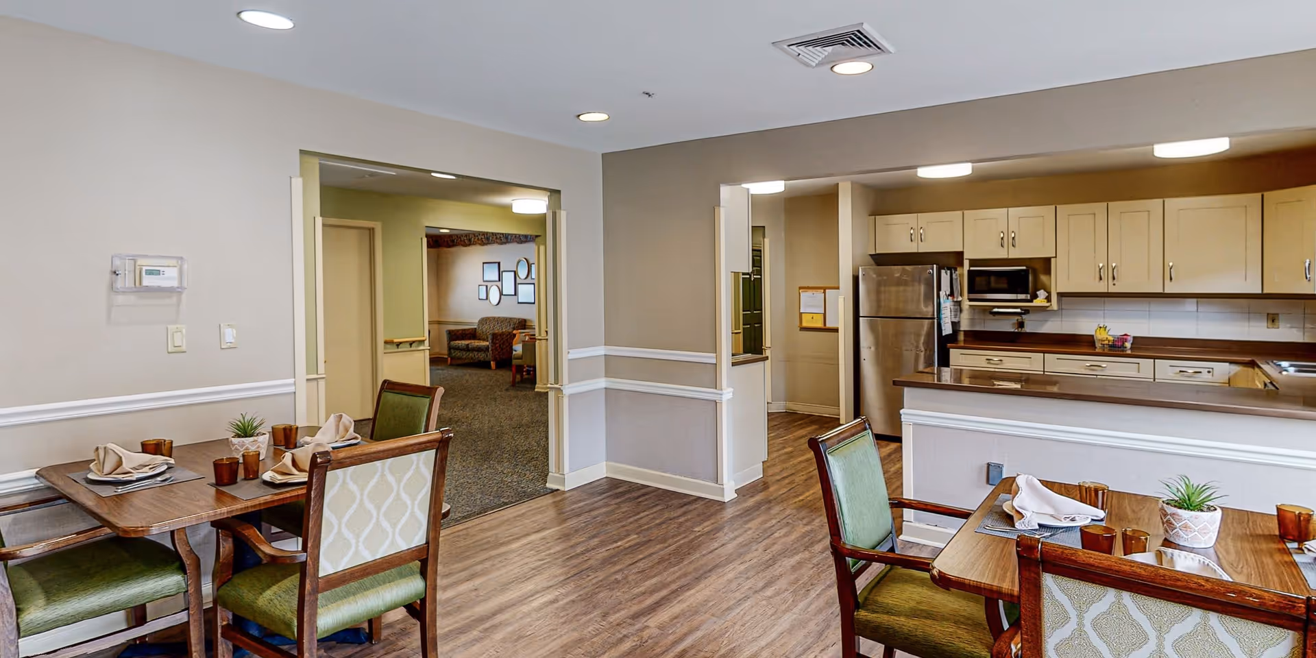 Interior view of a senior living facility showing a dining area with two wooden tables set with napkins, plates, and cups. The tables have green cushioned chairs. In the background, there is a kitchen with beige cabinets, a stainless steel refrigerator, and a microwave. Beyond the dining area, there is a living room with seating and framed pictures on the wall.
