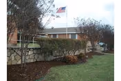 Exterior view of a brick building with a stone retaining wall, bushes, and an American flag on a flagpole in front of the building under a cloudy sky.