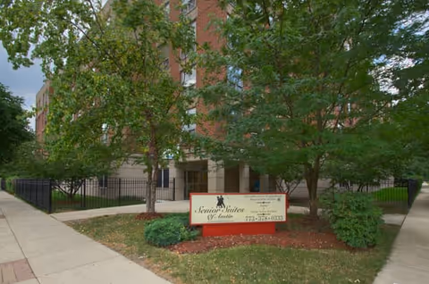 Front entrance of a brick senior living building with trees and a lawn sign reading 'Senior Suites' by the sidewalk.