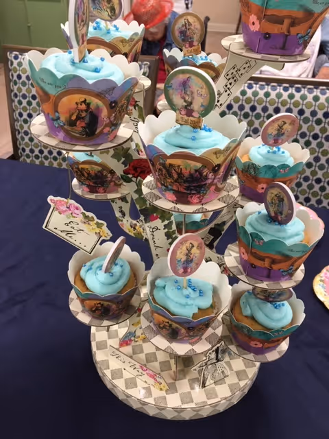 A multi-tiered cupcake stand holding several cupcakes with blue frosting and decorative toppers featuring vintage-style illustrations. The stand and cupcake wrappers have a whimsical, colorful design with various patterns and images. The setup is on a dark blue tablecloth with a patterned background.