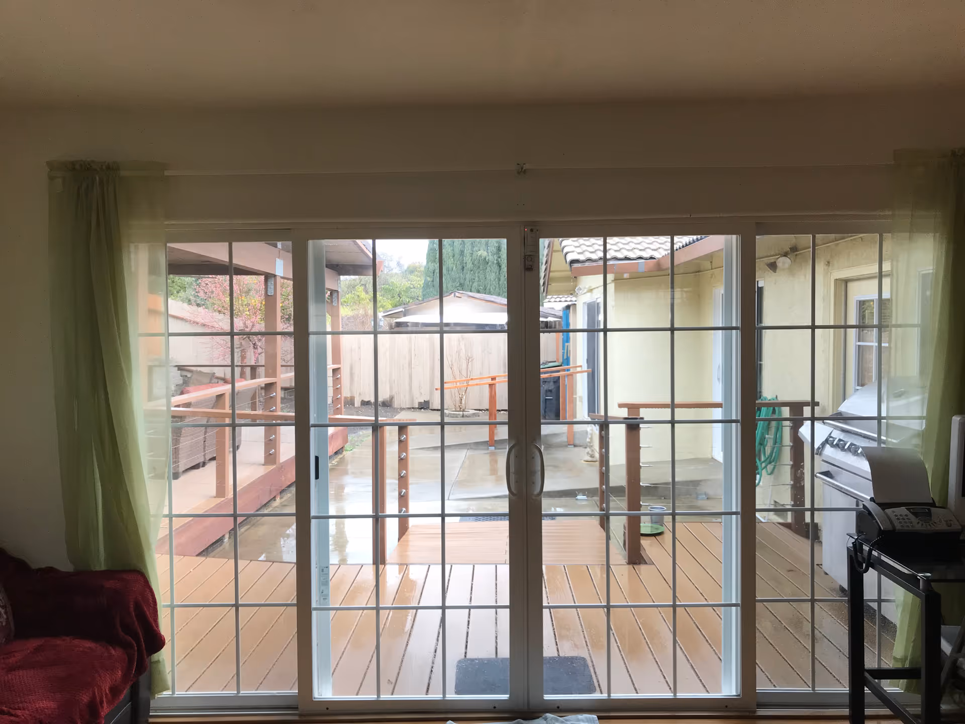 Sliding glass patio doors with green curtains open onto a wet wooden deck and backyard, viewed from a living room.