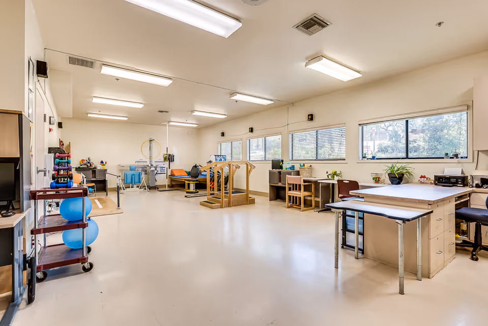 Bright therapy/activity room with exercise equipment, work tables, and large windows.