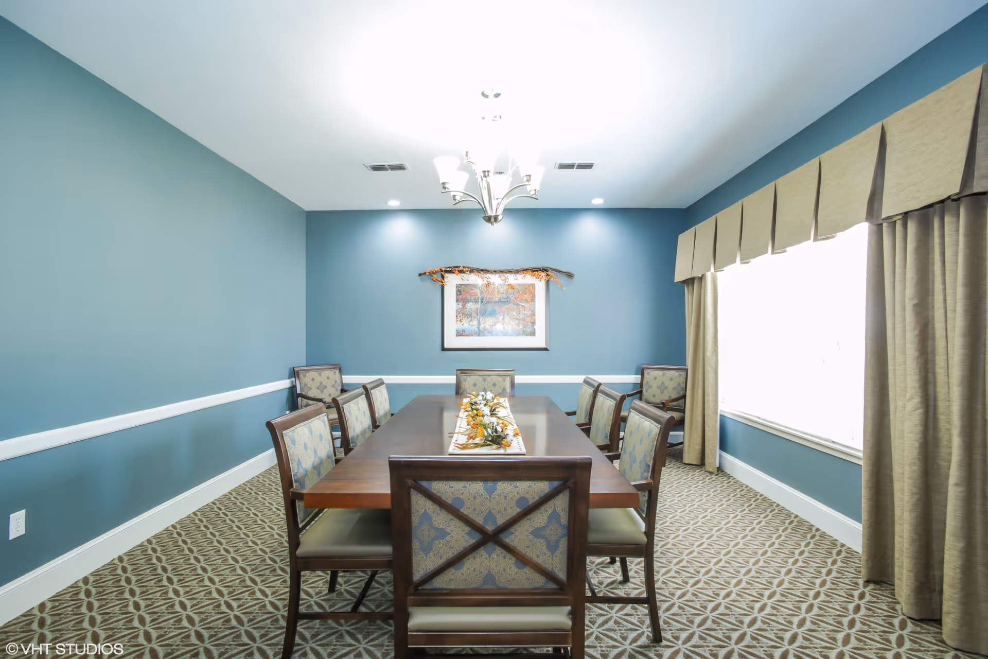 A dining room with a long wooden table surrounded by eight upholstered chairs. The walls are painted blue with white trim, and a framed artwork hangs on the far wall. A chandelier is mounted on the ceiling, and large windows with beige curtains allow natural light into the room. The floor is carpeted with a patterned design.