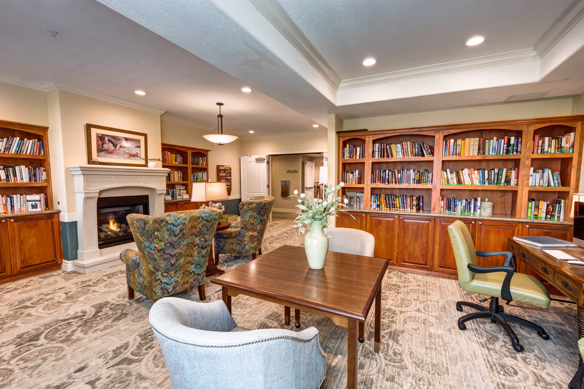 Well-lit senior living library/lounge with bookshelves, a fireplace, tables and upholstered chairs.