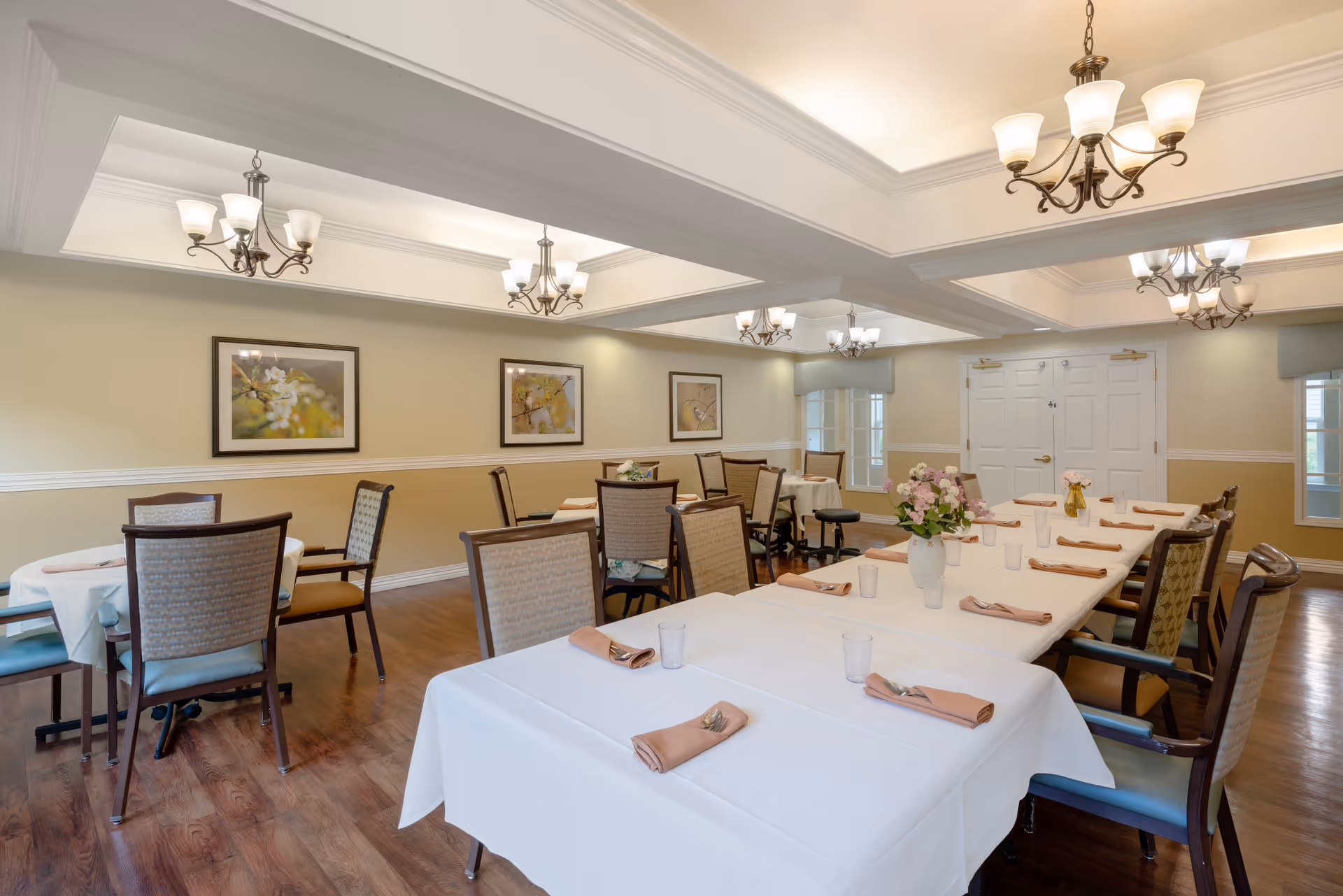 A well-lit dining room with multiple tables covered in white tablecloths, each set with folded beige napkins, utensils, and clear drinking glasses. The room features wooden chairs with patterned upholstery, hardwood floors, beige walls with white trim, three framed floral artworks, and elegant chandeliers hanging from a coffered ceiling. There are double white doors and windows with light blue valances in the background.