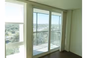 Sliding glass patio doors with vertical blinds opening onto a small balcony overlooking trees and distant buildings, viewed from an empty room with dark hardwood floors.