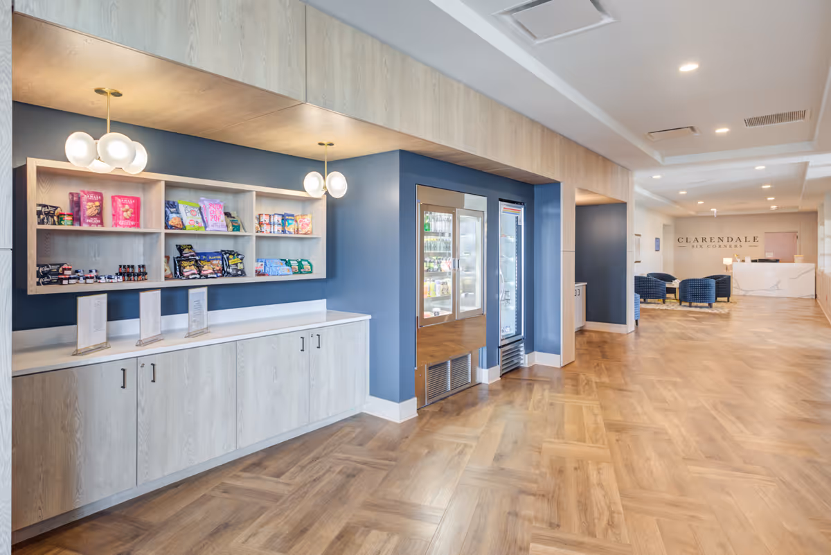 Interior view of a senior living facility lobby area with a snack and beverage station on the left featuring shelves stocked with snacks and refrigerated drinks. The floor has a wood pattern, and the walls are painted blue and white. In the background, there is a reception desk with the sign 'Clarendale Six Corners' on the wall and a seating area with blue chairs.