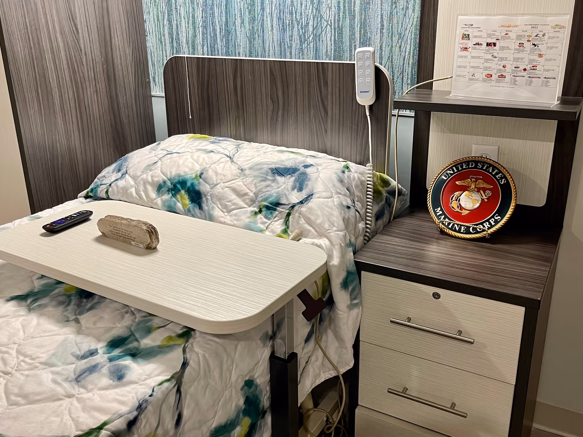 A hospital-style bed with a floral quilt, overbed table and remote next to a bedside cabinet displaying a United States Marine Corps plaque.