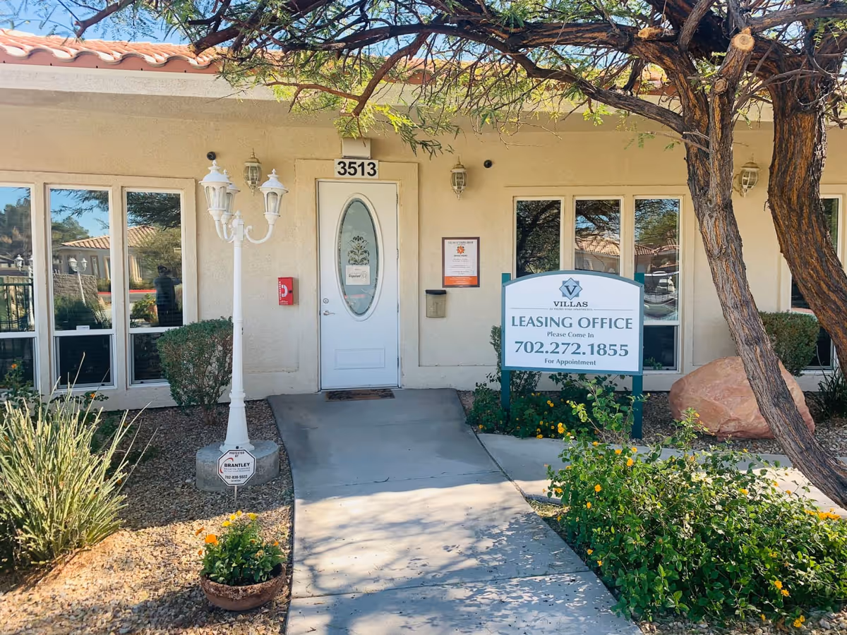 Exterior view of the leasing office entrance at The Villas at Viking Road, featuring a white door with an oval glass panel, a sign with leasing office information, surrounding bushes, a tree, and a white lamp post.