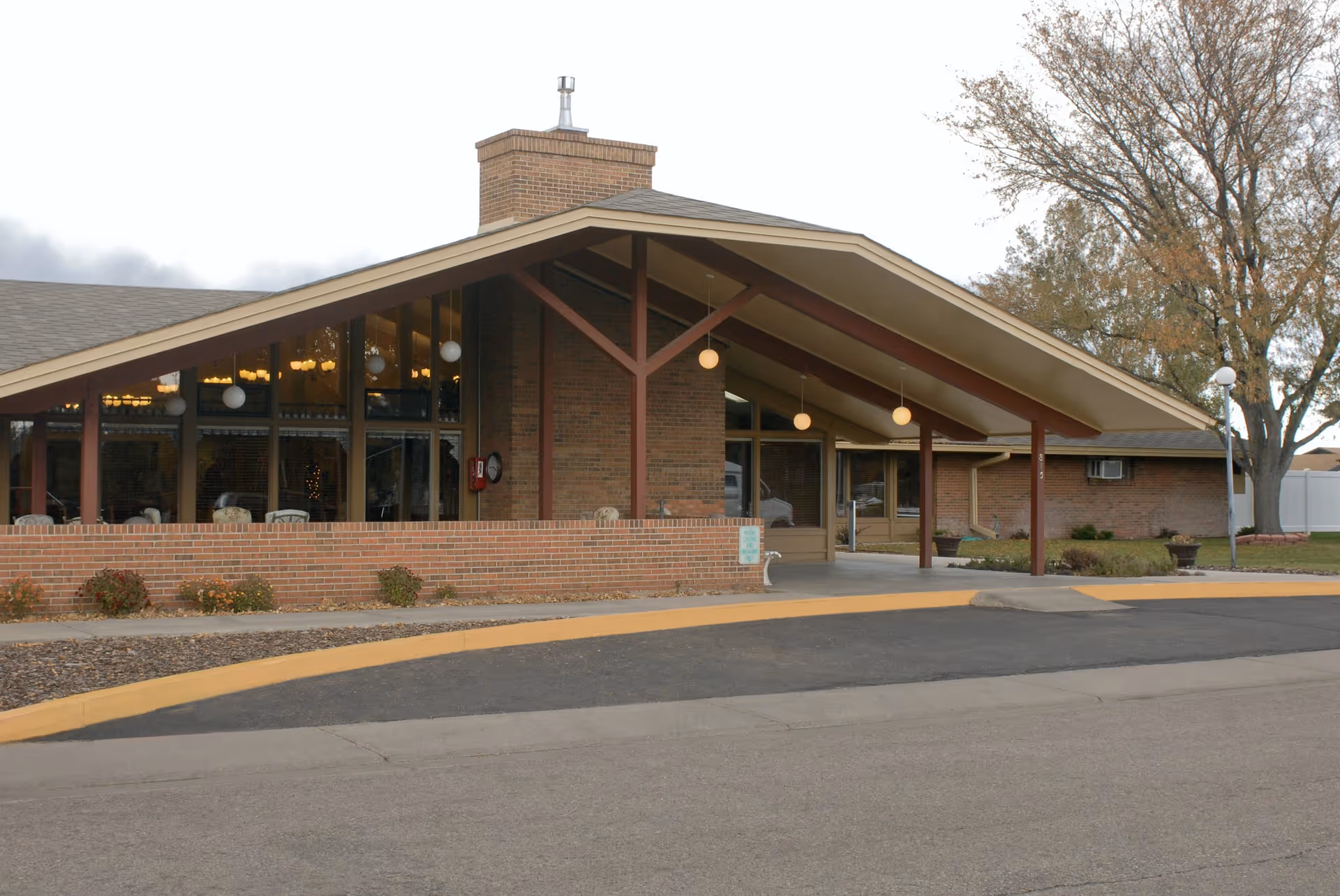 Exterior view of a single-story brick building with a large sloped roof and a covered entrance supported by beams. The building has large windows with visible interior lighting and a chimney. There are some shrubs and a tree nearby, and a paved driveway with a yellow curb in front.