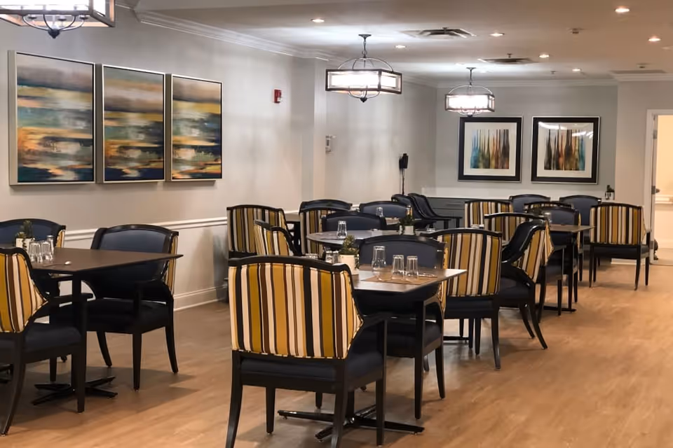 A dining room with multiple tables and chairs featuring striped upholstery. The room has wooden flooring, soft lighting from ceiling fixtures, and abstract artwork on the walls.
