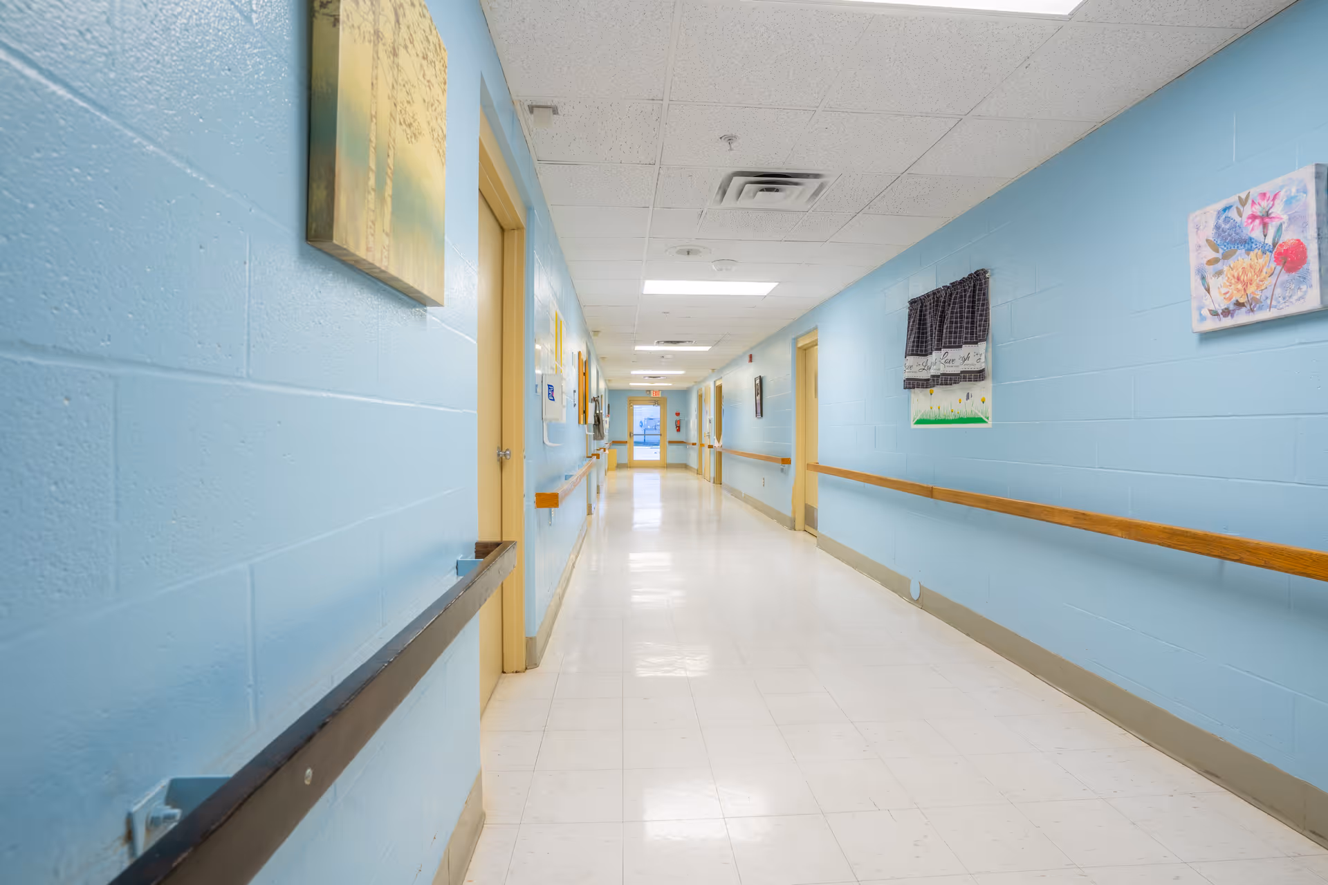 A long, clean hallway in a healthcare facility with light blue walls, white tiled floor, and a white ceiling with fluorescent lights. The hallway has wooden handrails on both sides and several closed doors along the walls. There are a few framed pictures and decorations hanging on the walls.