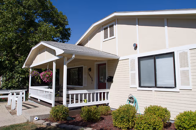 Exterior view of a beige and white single-story building with a covered porch, white railing, and a red door. There are green bushes and a garden hose near the building, with trees in the background under a clear blue sky.