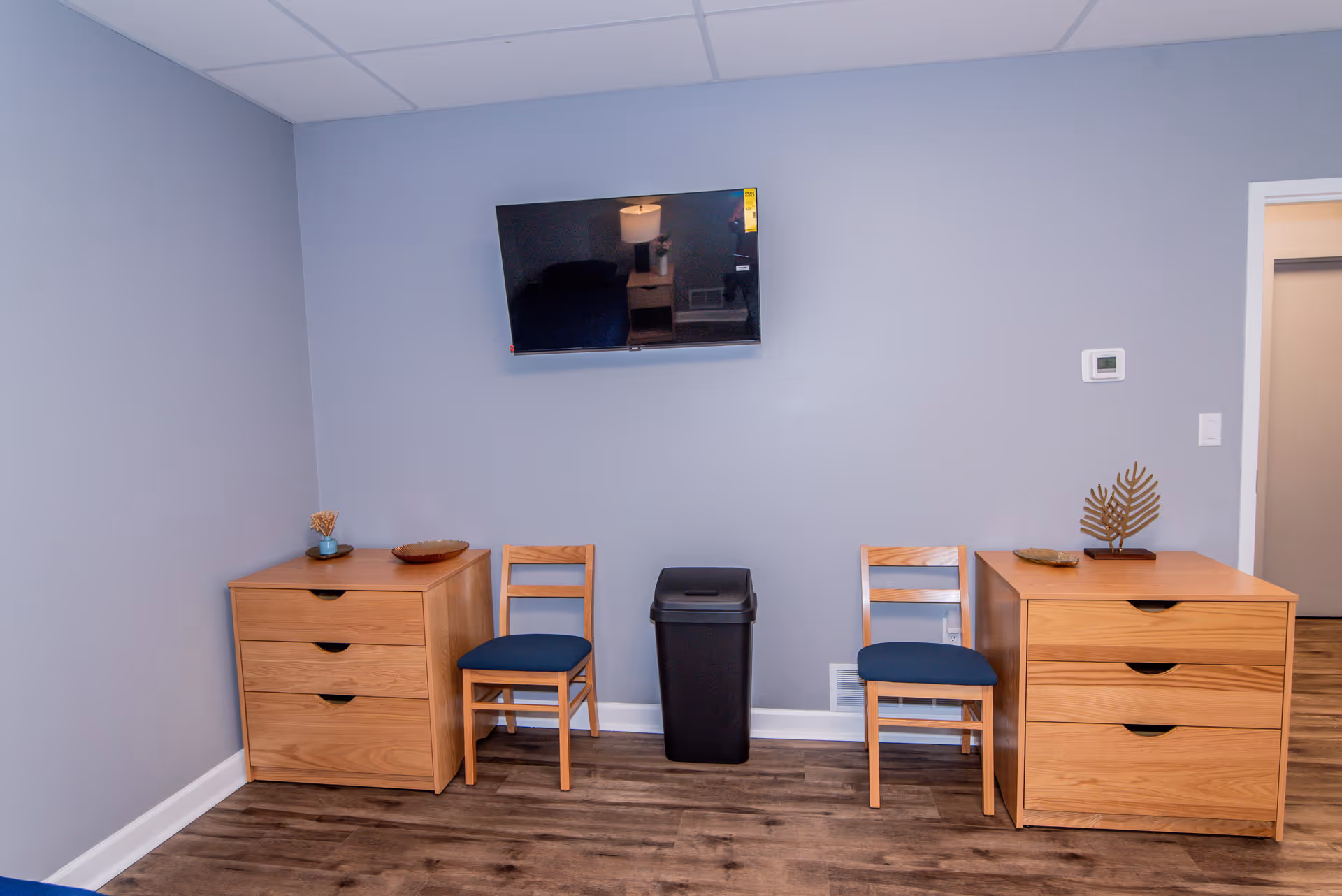 A room with two wooden dressers, each with three drawers, and two wooden chairs with blue cushions placed between them. A black trash bin is positioned between the chairs. A flat-screen TV is mounted on the light gray wall above the trash bin. The floor is wood-patterned, and there is a doorway visible on the right side of the image.